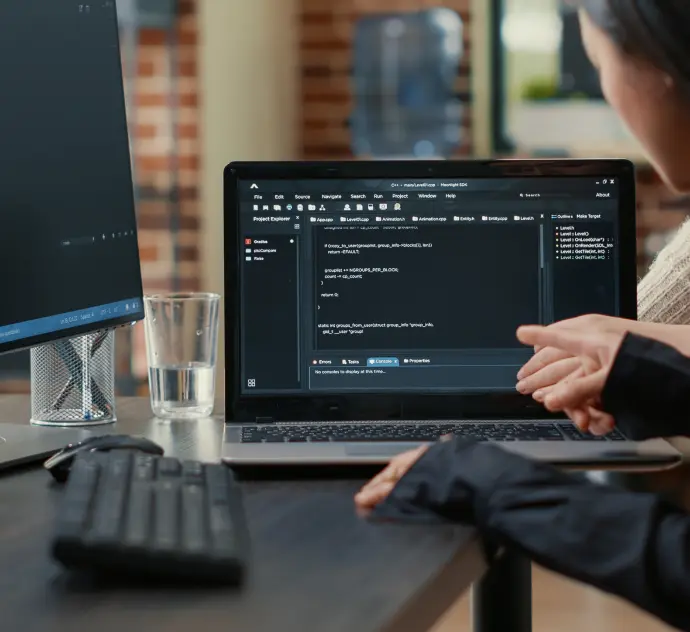 A person points at a laptop screen displaying code in a modern office. A glass of water and a monitor are on the desk. The atmosphere is focused and professional.