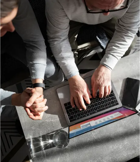 Two people are seated, viewed from above. One types on a laptop, while the other clasps their hands. A glass of water and a phone are nearby on a sunlit table.