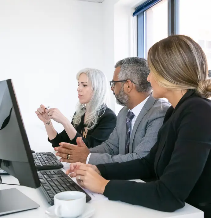 Three professionals in business attire sit at a desk, focused on a computer screen. They appear engaged and in discussion, with a cup of coffee nearby.