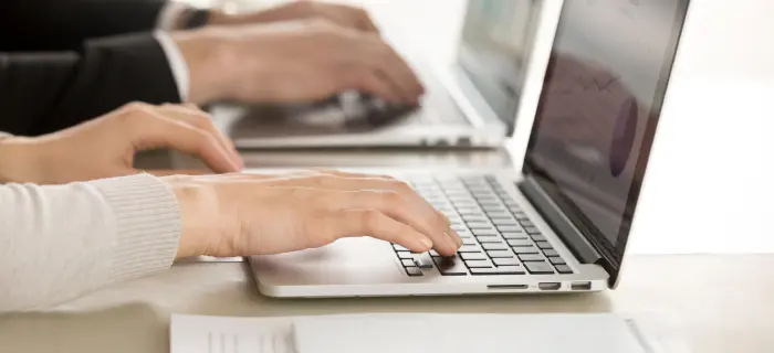 Hands typing on two laptops positioned side by side, suggesting teamwork or a collaborative work environment. The atmosphere is focused and professional.