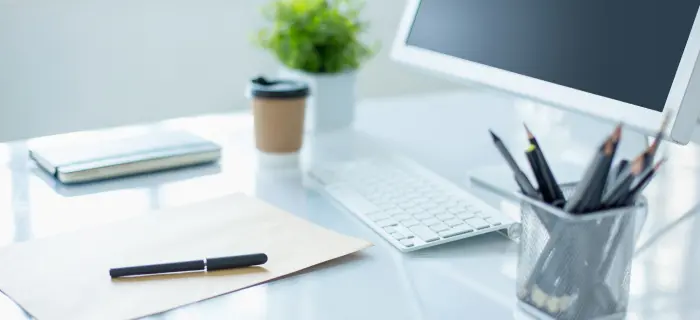A clean, modern desk setup with a computer monitor, white keyboard, pencil holder, coffee cup, notepad, and pen. A plant adds a touch of greenery.