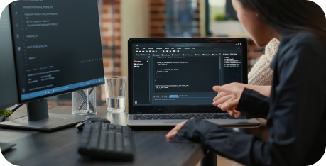 Two individuals collaborate at a desk with a laptop displaying code. A monitor and keyboard are nearby, conveying focus and teamwork.