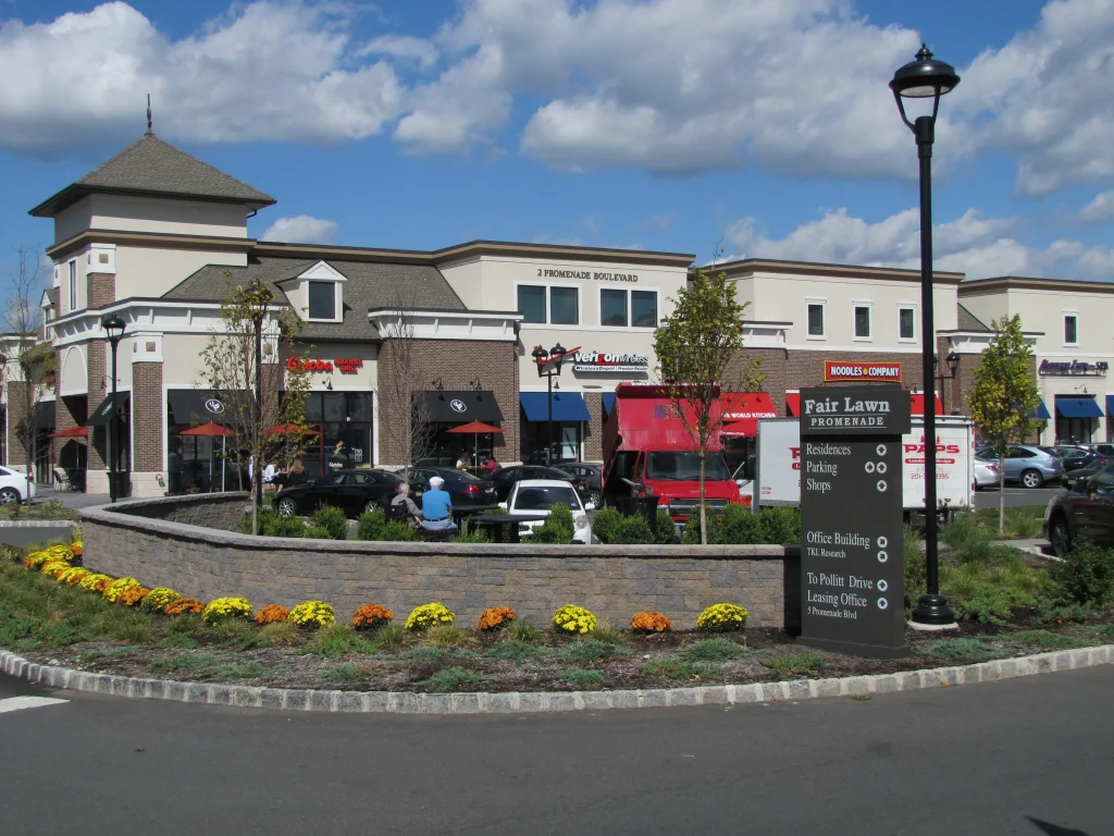 Shopping center with a brick facade and tower, featuring restaurants and shops. Colorful flowers in front. Blue sky and white clouds above.