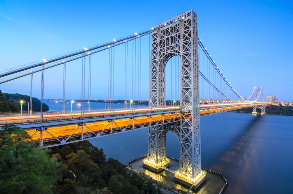 Suspension bridge illuminated at dusk, stretching over a calm river. The sky is a deep blue, and city lights are visible in the distance, creating a serene scene.