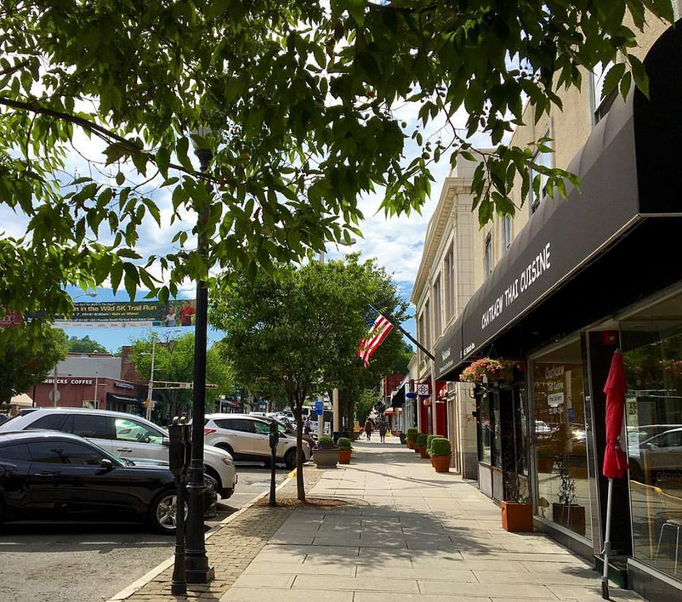 A sunny street scene with trees and cars lining a sidewalk. Shops, including a Thai restaurant, are on the right. An American flag hangs prominently, creating a lively and welcoming atmosphere.