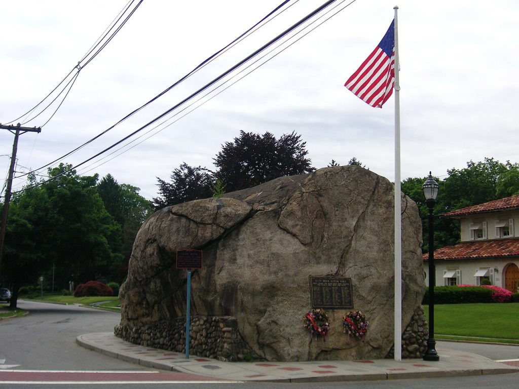 A large rock with commemorative plaques sits on a street corner, adorned with flowers. An American flag flies beside it, with overcast skies above.