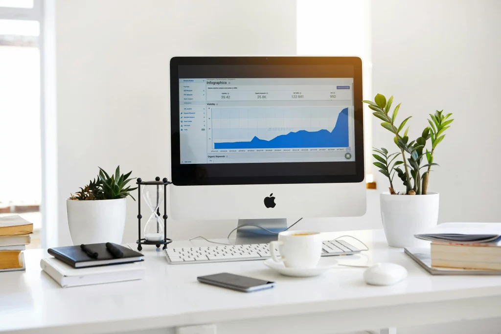 A minimalist office desk features a computer displaying a rising graph. Two potted plants, books, notebooks, and a coffee cup create a clean, focused workspace atmosphere.