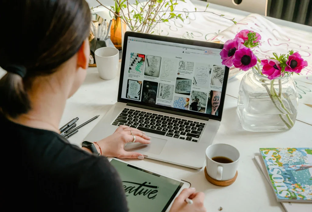 A woman working at a table, browsing art on a laptop, and using a tablet with a "creative" design. Bright flowers and a magazine add color and charm.