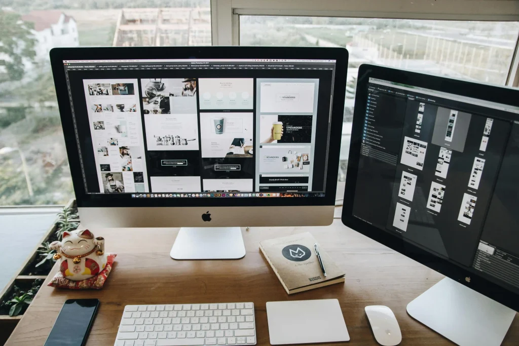 A modern desk setup with two monitors displaying web design projects. A white keyboard, mouse, smartphone, notebook, and a lucky cat figurine are on the wooden desk.