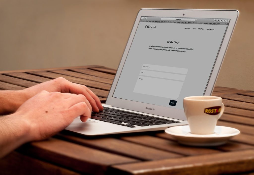 Hands typing on a laptop at a wooden table with a contact form open. A coffee cup marked "ROST" sits on a saucer, suggesting a relaxed, focused work setting.