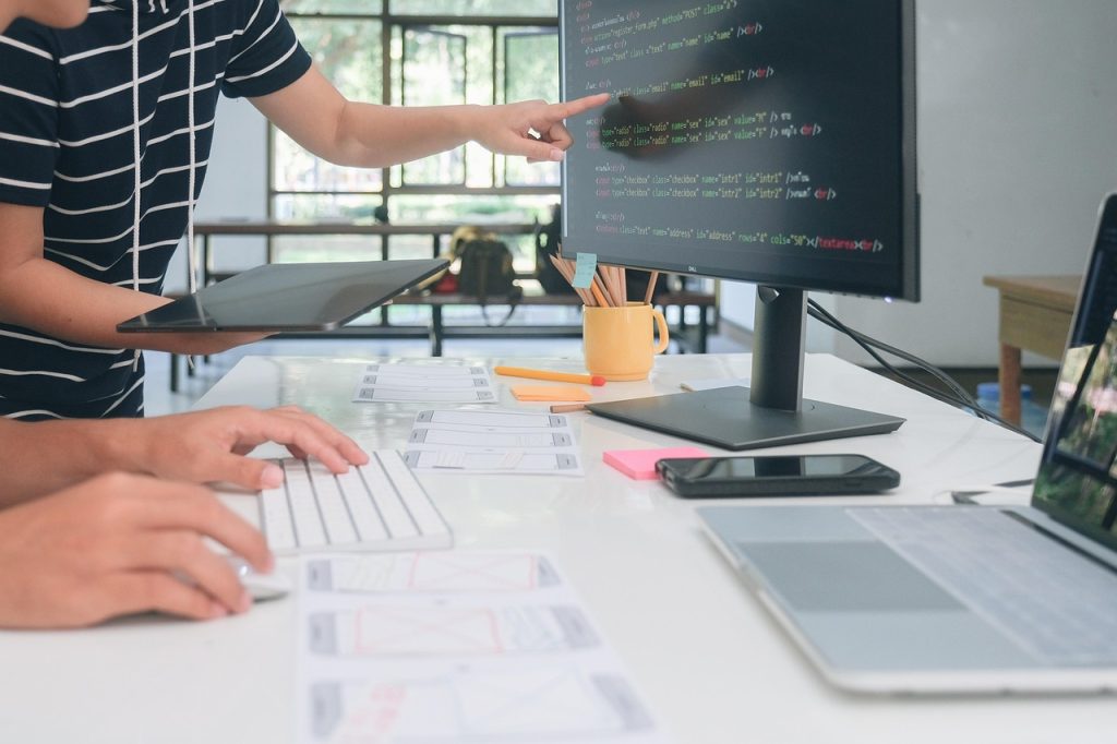 Two people collaborate at a desk with a computer monitor displaying code. One types on a keyboard, while the other points at the screen. The scene conveys teamwork.