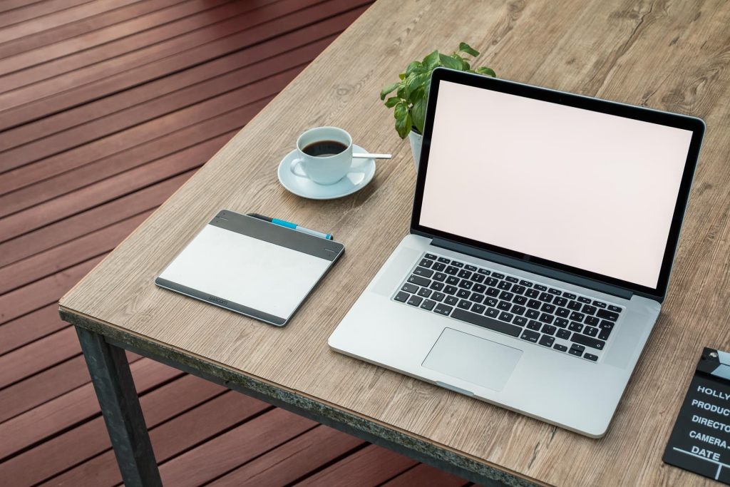 A laptop with a blank screen on a wooden table, next to a graphics tablet, a cup of coffee, and a small plant. The setting is outdoors on a deck.
