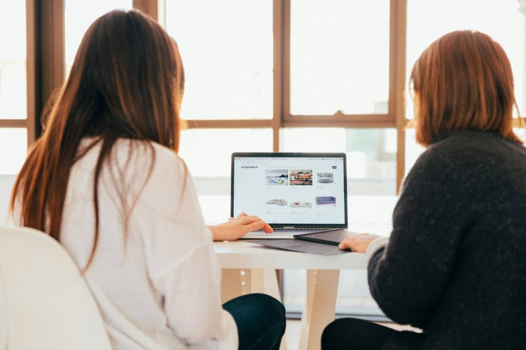 Two people seated at a table, viewing a laptop screen displaying a website. They appear engaged in discussion, with bright windows in the background.